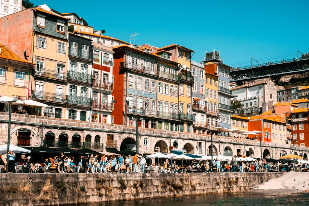 Colorful buildings line the waterfront of Ribeira, Porto, with people strolling along the Douro River under vibrant blue skies.